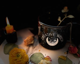 May include: Black ceramic mug with a white crescent moon and stars design. The mug says "Creative Witch" and has a gold chain handle. The mug is sitting on a white marble surface with dried flowers and leaves.