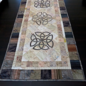 Pode incluir: Um corredor de mesa bege e cinza com um design de nó celta. O corredor é feito de tecido patchwork com um padrão floral.
