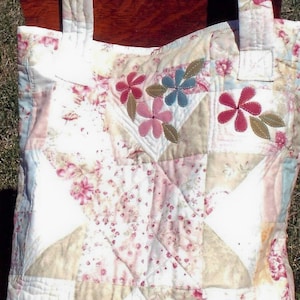 Op de afbeelding: Een witte gewatteerde tote bag met bloemenpatchwork en aangebrachte bloemen in roze, blauwe en bruine tinten. De tas heeft twee hengsels.