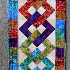 May include: A colourful patchwork quilt runner with a geometric pattern of squares and diamonds. The runner features a variety of colours, including red, orange, yellow, green, blue, purple, and white.
