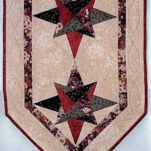 May include: A quilted table runner featuring two large starburst designs. The stars are composed of red, black, and gray geometric shapes with floral accents. The runner has a cream background with a floral border and a burgundy edge.