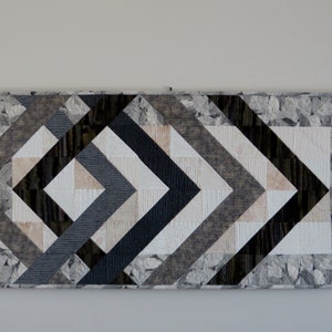 May include: A geometric table runner with a modern design. The runner is made of fabric in shades of gray, black, and beige. The fabric has a subtle pattern of leaves.
