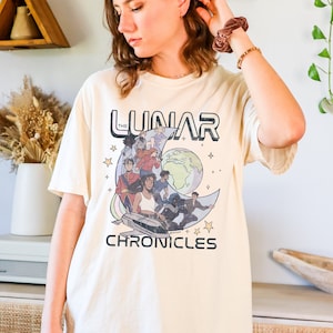 Può includere: Una t-shirt color crema con una stampa grafica di una luna crescente con un'immagine di cartoni animati di un gruppo di persone di fronte a un pianeta. Il testo "The Lunar Chronicles" è stampato sotto l'immagine.