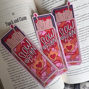 Three "Slow Burn" matchbox-shaped bookmarks with a pink and red design, featuring flames and hearts. The bookmarks are placed on an open book. The text "Made for Readers" is also visible.
