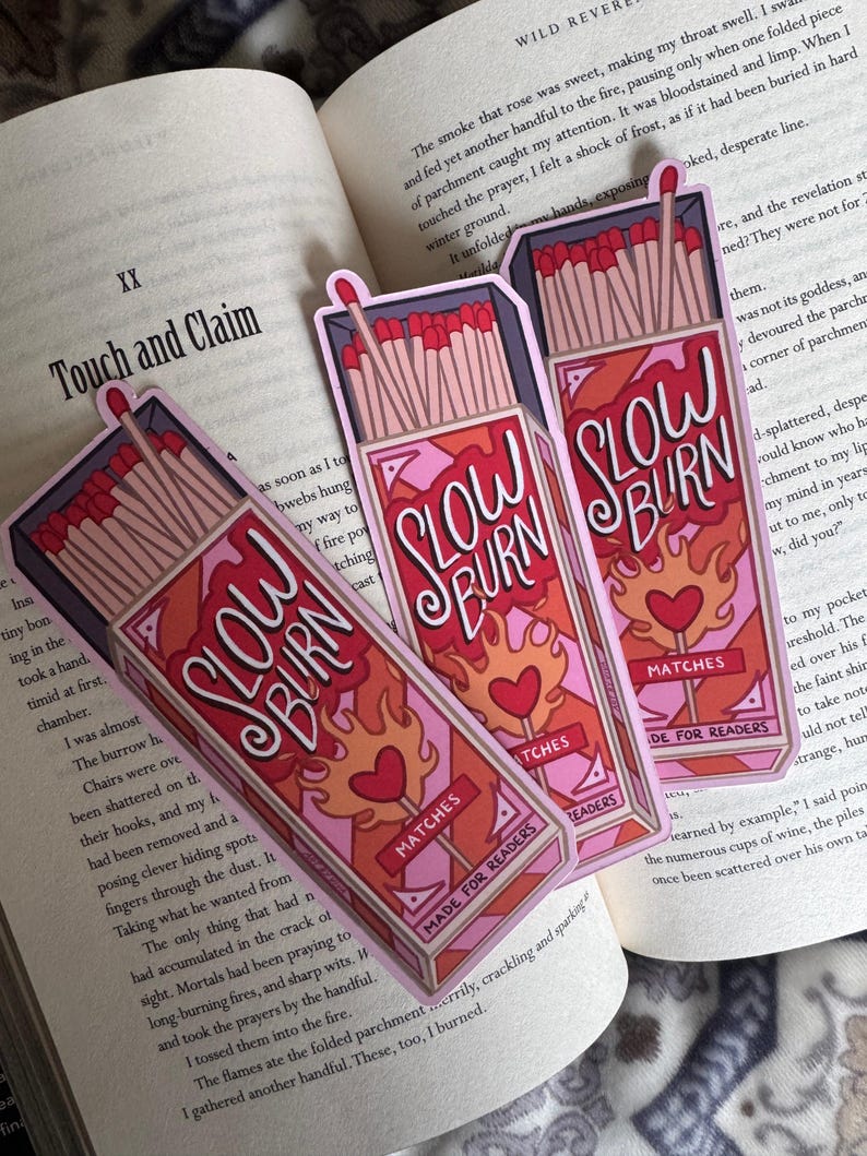 Three "Slow Burn" matchbox-shaped bookmarks with a pink and red design, featuring flames and hearts. The bookmarks are placed on an open book. The text "Made for Readers" is also visible.