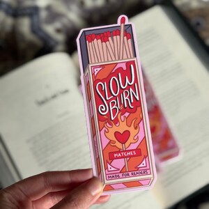 A bookmark featuring a matchbox design with the words "SLOW BURN" in red and white. The matchbox has a heart-shaped flame design and the text "MATCHES MADE FOR READERS". The sticker is held over an open book.