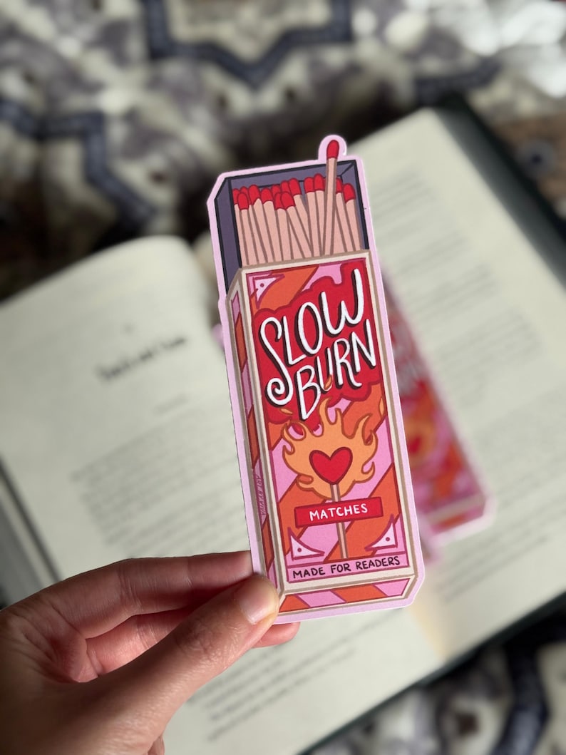 A bookmark featuring a matchbox design with the words "SLOW BURN" in red and white. The matchbox has a heart-shaped flame design and the text "MATCHES MADE FOR READERS". The sticker is held over an open book.