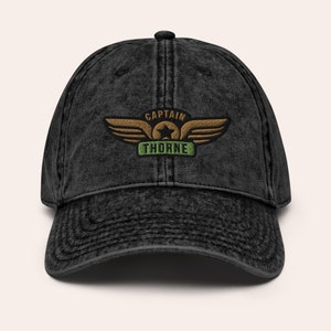 May include: Black distressed denim baseball cap with an embroidered patch that says "Captain Thorne" with a star and wings.