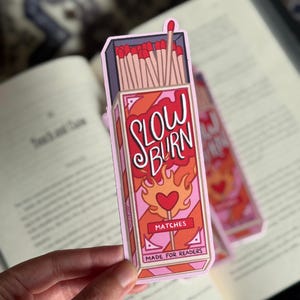 A bookmark featuring a matchbox design with the words "SLOW BURN" in red and white. The matchbox has a pink and orange color scheme with a heart and flames graphic. The text "MATCHES" and "MADE FOR READERS" are also visible.