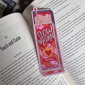 A pink and red bookmark in the shape of a matchbox, with the words "SLOW BURN" in a stylized font. The bookmark also features a heart and flames design, with the word "MATCHES" and "MADE FOR READERS" below.