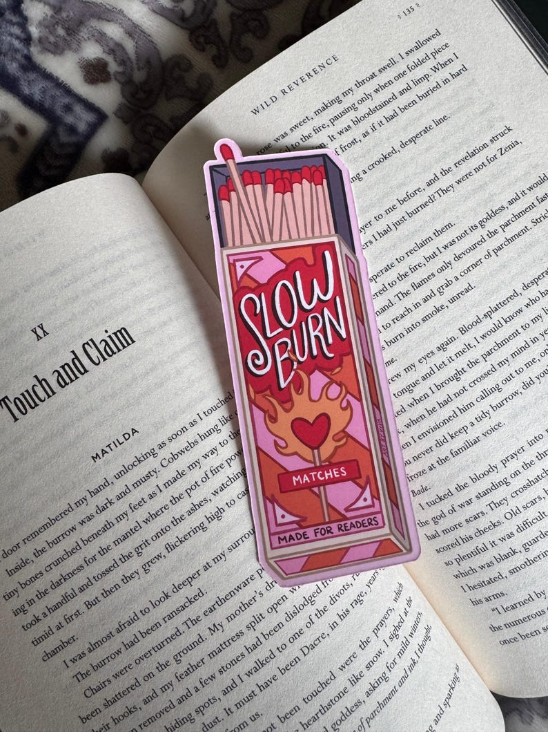 A pink and red bookmark in the shape of a matchbox, with the words "SLOW BURN" in a stylized font. The bookmark also features a heart and flames design, with the word "MATCHES" and "MADE FOR READERS" below.