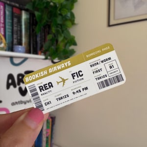 Peut inclure: Un billet d'embarquement fantaisie avec le texte "BOOKISH AIRWAYS" et les destinations "REA" à "FIC". Le billet comprend les détails du vol : TBR125, 21h45, et les informations du passager : BOOK/WORM, première classe, siège A1. Le design est en blanc, or et noir.
