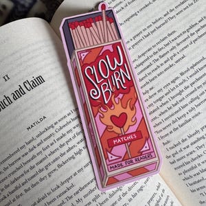 A rectangular bookmark featuring a stylized matchbox design. The bookmark is predominantly pink and red, with the words "SLOW BURN" in a handwritten font. The design includes a heart and the text "MATCHES MADE FOR READERS".