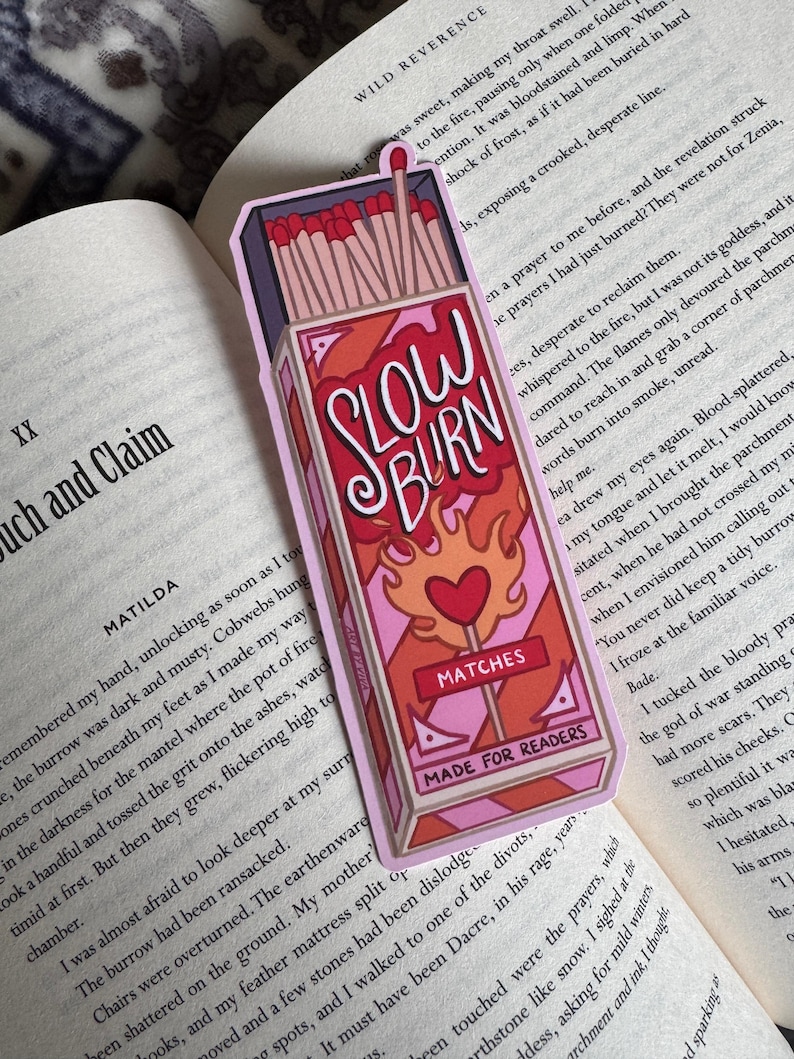 A rectangular bookmark featuring a stylized matchbox design. The bookmark is predominantly pink and red, with the words "SLOW BURN" in a handwritten font. The design includes a heart and the text "MATCHES MADE FOR READERS".