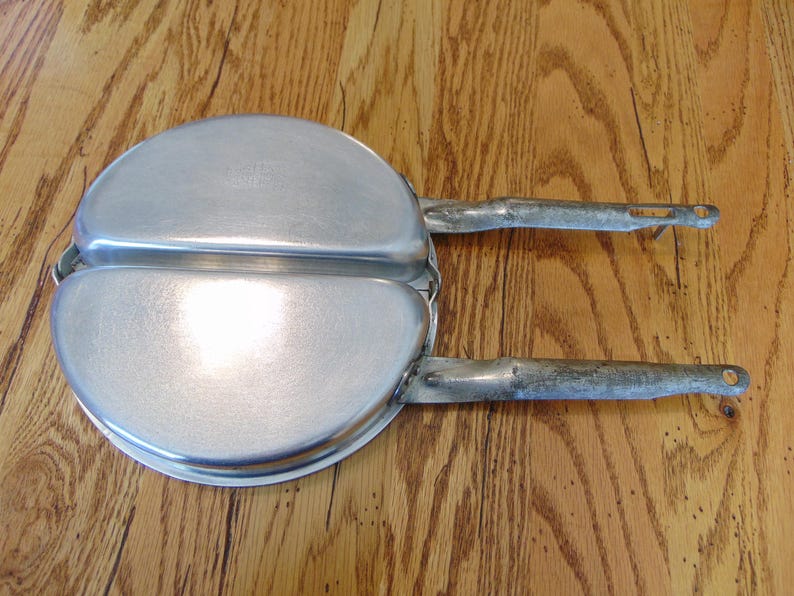 Puede incluir: Una sart&eacute;n de camping vintage de dos piezas, de color plateado, con dos asas. La sart&eacute;n es de metal y tiene una superficie ligeramente desgastada.