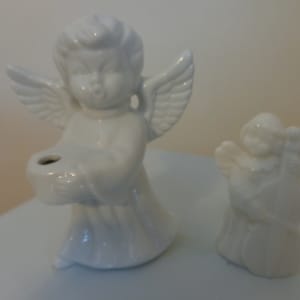 May include: Two white porcelain angel figurines. One angel is holding a small, round container, while the other angel is holding a musical instrument.