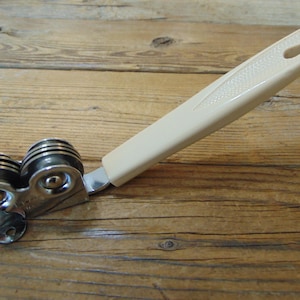 May include: A vintage can opener with a cream-colored handle and a dual-wheel design. The handle has a textured pattern and a hole for hanging. The opener's metal components are silver-toned.