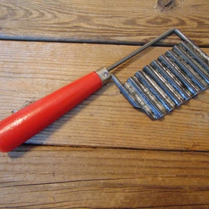 May include: A vintage crinkle cutter with a bright red wooden handle. The metal blade has a wavy design for creating decorative cuts. The cutter is designed for use in the kitchen for cutting vegetables and fruits.