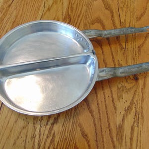 Puede incluir: Una sart&eacute;n de camping vintage de aluminio dividida con dos asas. La sart&eacute;n est&aacute; dividida en dos y tiene una superficie lisa y brillante.
