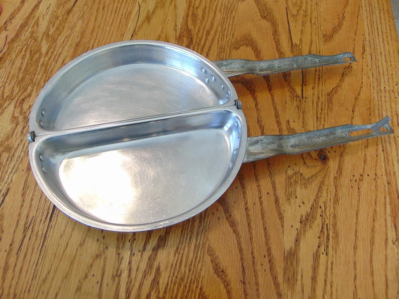 Puede incluir: Una sart&eacute;n de camping vintage de aluminio dividida con dos asas. La sart&eacute;n est&aacute; dividida en dos y tiene una superficie lisa y brillante.