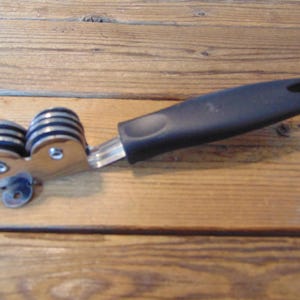 May include: A knife sharpener with a black handle and two cylindrical sharpening wheels. The wheels are silver and black, mounted on a silver metal frame. The handle has a hole for hanging.