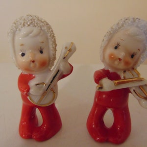 May include: Two vintage porcelain figurines of children wearing red outfits and white hats. One child is playing a banjo and the other is playing a violin.