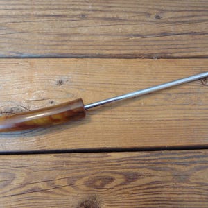May include: A vintage fondue fork with a stainless steel shaft and a two-pronged end. The handle is made of a brown and yellow marbled material. The fork is lying on a wooden surface.