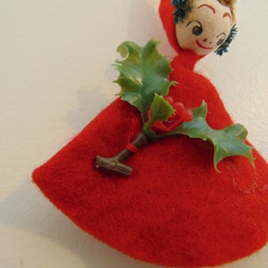 May include: A red felt Christmas ornament with a painted face and a green holly sprig with red berries.