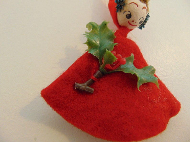 May include: A red felt Christmas ornament with a painted face and a green holly sprig with red berries.