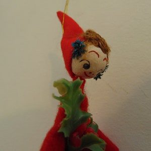 May include: A vintage Christmas ornament featuring a felt elf with a red hat, brown hair, and a painted face. The elf is holding a green holly sprig.