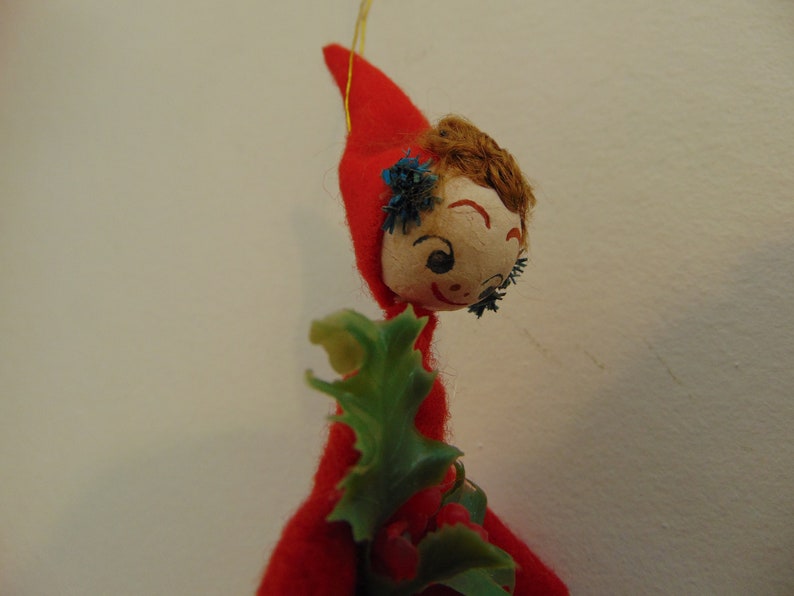 May include: A vintage Christmas ornament featuring a felt elf with a red hat, brown hair, and a painted face. The elf is holding a green holly sprig.