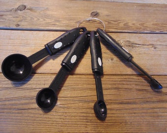 May include: A set of five black measuring spoons with white markings on the handles. The spoons are connected by a metal ring and are arranged in a fan shape on a wooden surface.