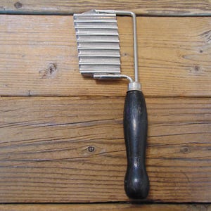 May include: A vintage crinkle cutter with a stainless steel blade and a dark wooden handle. The blade has a wavy design for creating crinkle-cut vegetables. The handle is curved for a comfortable grip. The cutter is set against a wooden background.