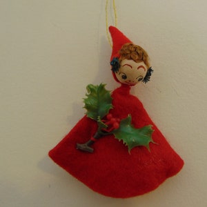 May include: A red felt Christmas ornament shaped like a girl wearing a red dress and holding a sprig of holly.