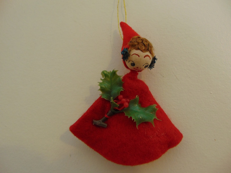 May include: A red felt Christmas ornament shaped like a girl wearing a red dress and holding a sprig of holly.