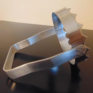 May include: A silver-colored metal tool with a jagged edge and a curved handle. The tool is designed with a claw-like shape, and is likely used for a specific task. The metal has a brushed finish, and the tool is set against a dark surface.