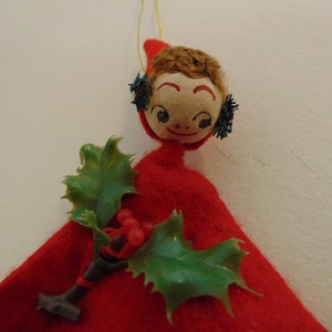 May include: A red felt Christmas ornament with a painted face, brown hair, and blue glitter accents. The ornament is holding a sprig of green holly with red berries.