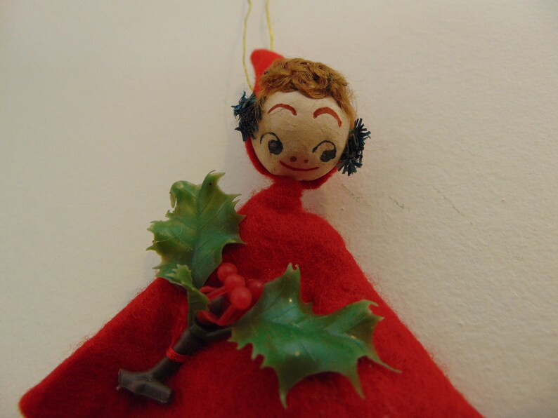 May include: A red felt Christmas ornament with a painted face, brown hair, and blue glitter accents. The ornament is holding a sprig of green holly with red berries.
