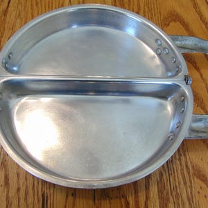 Puede incluir: Un plato de camping dividido de aluminio plateado con un asa con bisagra. El plato est&aacute; dividido en dos secciones y tiene un peque&ntilde;o borde alrededor del borde.