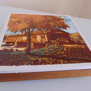 May include: A wooden framed tile with a colorful image of a covered bridge in a fall setting. The bridge is brown with a red roof and is surrounded by trees with orange and yellow leaves. The tile is a great addition to any home decor.