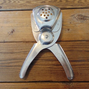 May include: A vintage, silver-toned citrus juicer. The juicer has a perforated top, a hinged design, and handles for squeezing. The tool is made of metal and is designed for pressing citrus fruits. The juicer is shown on a wooden surface.