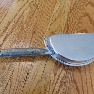 Puede incluir: Una sart&eacute;n de camping de metal plateado con un mango largo y delgado. El mango est&aacute; unido a la sart&eacute;n con una bisagra de metal.