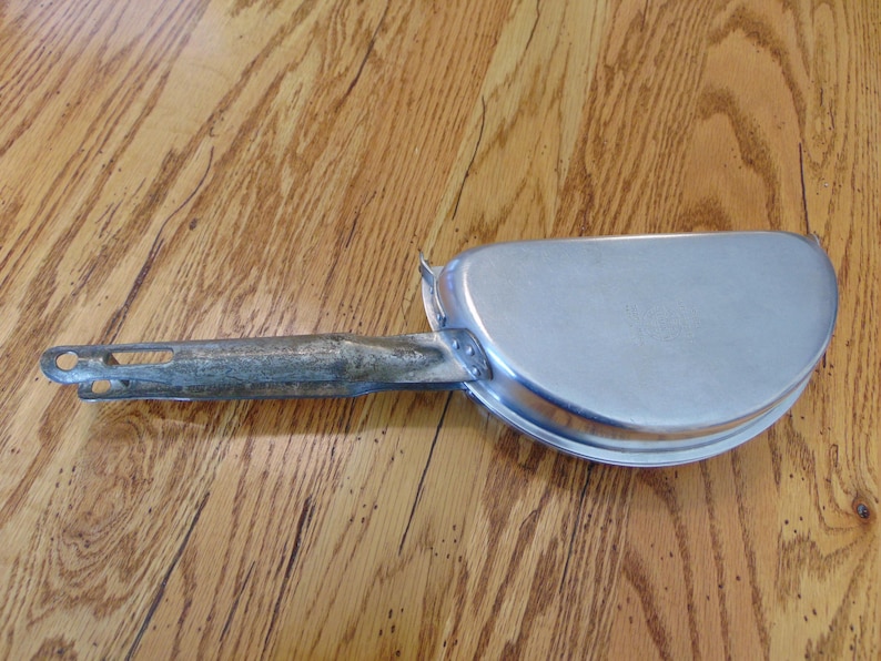 Puede incluir: Una sart&eacute;n de camping de metal plateado con un mango largo y delgado. El mango est&aacute; unido a la sart&eacute;n con una bisagra de metal.