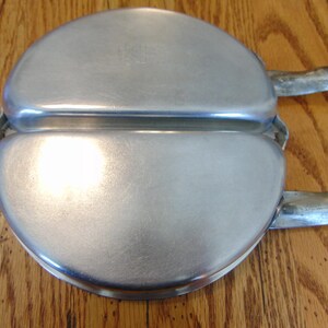 Puede incluir: Una plancha para gofres de metal plateado con dos secciones redondas y dos asas. La plancha para gofres est&aacute; cerrada.