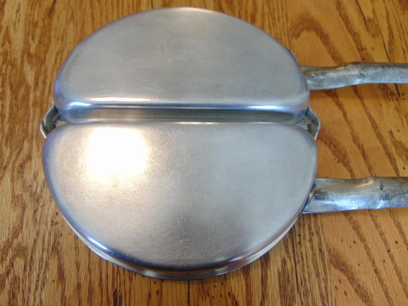 Puede incluir: Una plancha para gofres de metal plateado con dos secciones redondas y dos asas. La plancha para gofres est&aacute; cerrada.