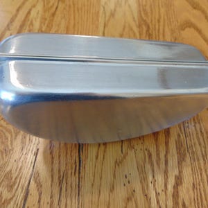 Puede incluir: Un recipiente de metal plateado con una tapa con bisagras. El recipiente tiene una superficie lisa y brillante y una forma redondeada.