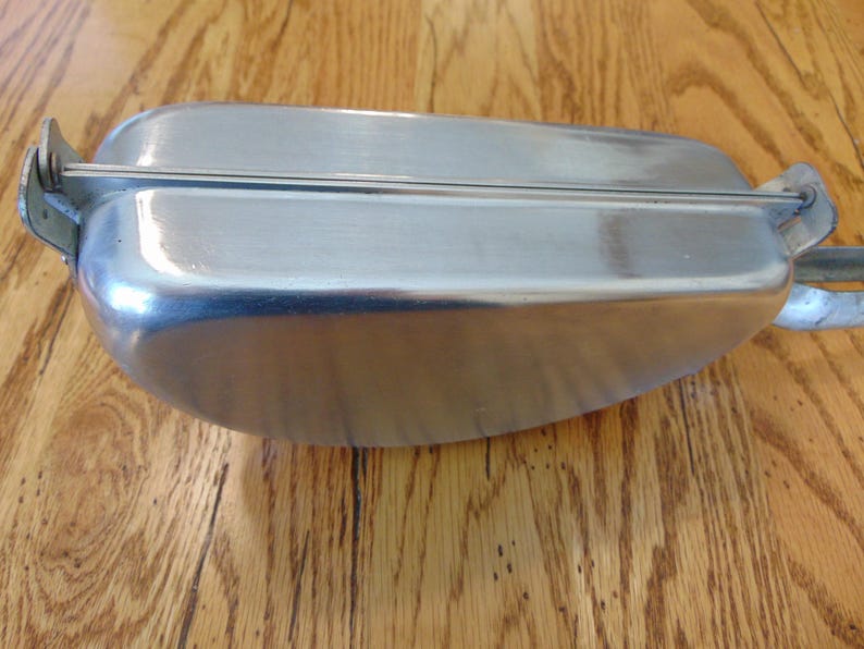 Puede incluir: Un recipiente de metal plateado con una tapa con bisagras. El recipiente tiene una superficie lisa y brillante y una forma redondeada.