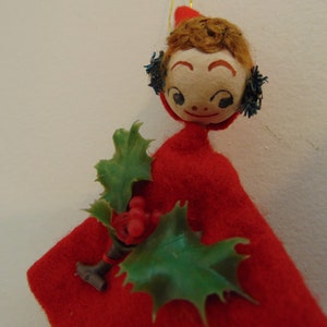 May include: A vintage felt Christmas ornament depicting a girl wearing a red dress and a holly sprig. The girl has a painted face with brown hair and blue eyes.