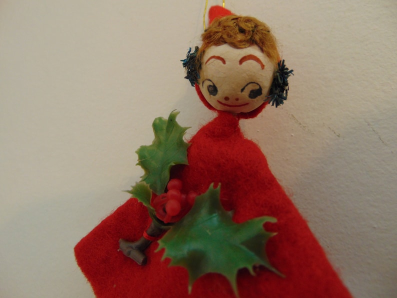 May include: A vintage felt Christmas ornament depicting a girl wearing a red dress and a holly sprig. The girl has a painted face with brown hair and blue eyes.