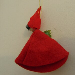 May include: A red felt Christmas ornament shaped like a person wearing a red pointed hat and a green holly sprig.